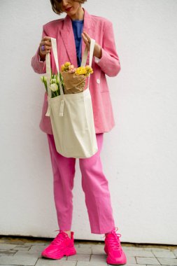 Stylish woman in pink suit with eco handbag and flowers on white background. Canvas bag with blank space