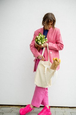 Stylish woman in pink suit takes some magazine from eco handbag and holds flowers on white background. Canvas bag with blank space