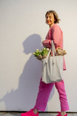 Stylish woman in pink suit walks with eco handbag full of flowers on white background. Canvas bag with blank space. Concept of style and spring time