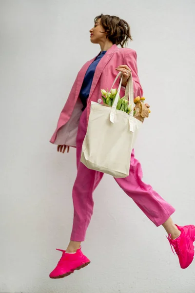 穿着粉红西服的时髦女人拿着装满白色背景花朵的生态手提包跳了起来 有空隙的帆布袋 — 图库照片