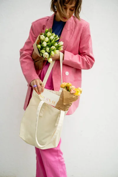 穿着粉色西服的时髦女人从环保手提包里拿出一本杂志 把花放在白色的背景上 有空隙的帆布袋 — 图库照片