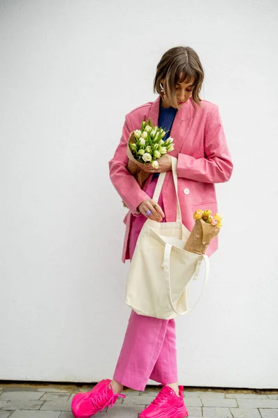 ピンクのスーツのスタイリッシュな女性は エコハンドバッグからいくつかの雑誌を取り 白い背景に花を保持しています 空白のキャンバスバッグ — ストック写真