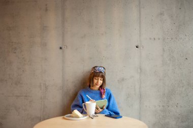 Young stylish woman works on a digital tablet while sitting on concrete wall background at modern coffee shop. Concept of remote creative work online