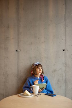 Young stylish woman works on a digital tablet while sitting on concrete wall background at modern coffee shop. Concept of remote creative work online