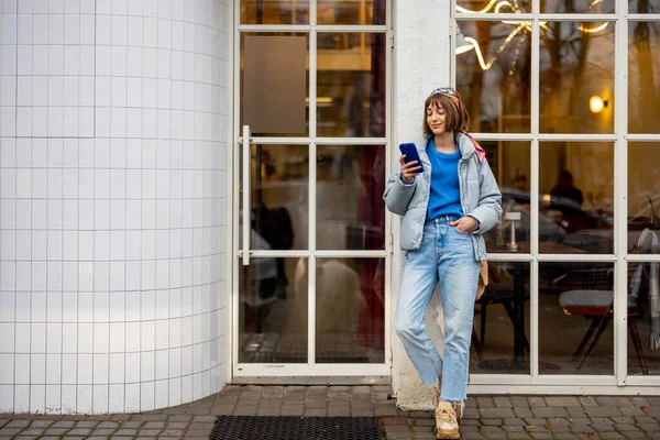 Young stylish woman in blue casual clothes stands with phone near modern cafe window outdoors. Concept of city lifestyle and style