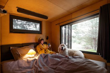 Woman uses phone while lying in bed with her cute dog in tiny bedroom of wooden cabin on nature