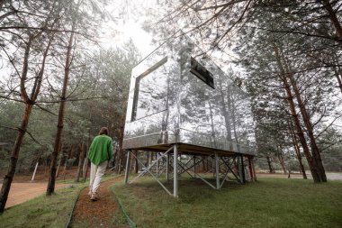 Woman near an invisible house with mirrored walls in pine forest. Connection with nature and sustainability concept. Rest in tiny cabins on nature