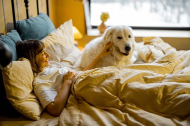 Woman Lying with her huge and cute dog in bed at cozy bedroom in beige tones. Concept of home coziness and love with pets