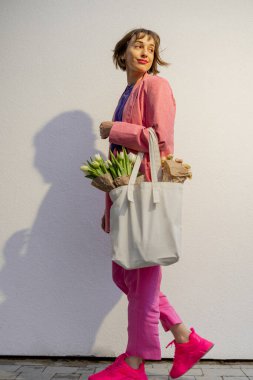 Stylish woman in pink suit walks with eco handbag full of flowers on white background. Canvas bag with blank space. Concept of style and spring time