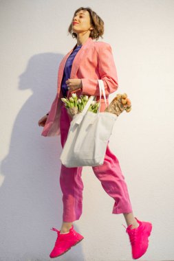 Stylish woman in pink suit jumps with eco handbag full of flowers on white background. Canvas bag with blank space. Concept of style and spring time