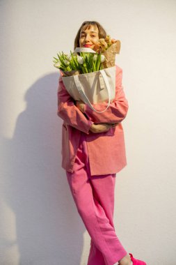 Stylish woman in pink suit carries eco handbag full of flowers on white background. Canvas bag with blank space. Concept of style and spring time