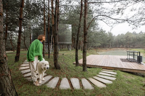 Woman walks on wavy pathway near lake and invisible cabin in pine forest. Connection with nature and sustainability concept. Rest in tiny cabins on nature