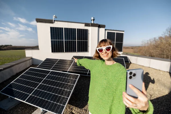 Young woman talks on phone while standing on rooftop with a solar power plant. Happy owner of energy-independent household. Eco-friendly and sustainable lifestyle