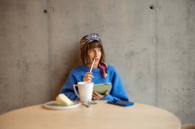 Young stylish woman works on a digital tablet while sitting on concrete wall background at modern coffee shop. Concept of remote creative work online