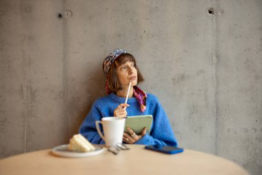 Young stylish woman works on a digital tablet while sitting on concrete wall background at modern coffee shop. Concept of remote creative work online