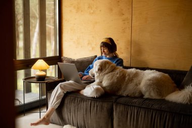 Young woman works on laptop while sitting on a couch with her white adorable dog at home. Concept of remote work from home and friendship with pets