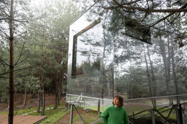 Woman near an invisible house with mirrored walls in pine forest. Connection with nature and sustainability concept. Rest in tiny cabins on nature