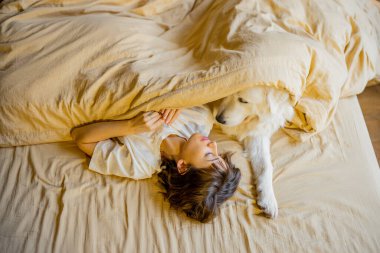 Young woman hugs with her cute dog while lying together covered with beige blanket in bed. View from above. Concept of friendship with pets and home coziness