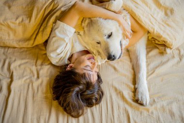 Young woman hugs with her cute dog while lying together covered with beige blanket in bed. View from above. Concept of friendship with pets and home coziness