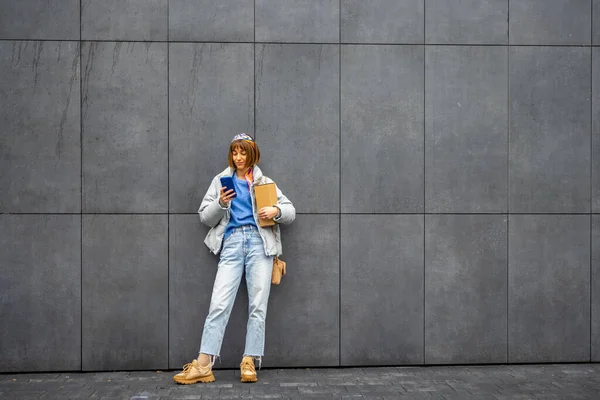 若いスタイリッシュな女性は 屋外の灰色の壁の背景に立っている間 携帯電話を使用しています コピースペース付きのワイドビュー — ストック写真