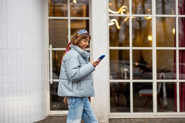 Young stylish woman in blue casual clothes walks with phone on background of modern cafe window outdoors. Concept of city lifestyle and style