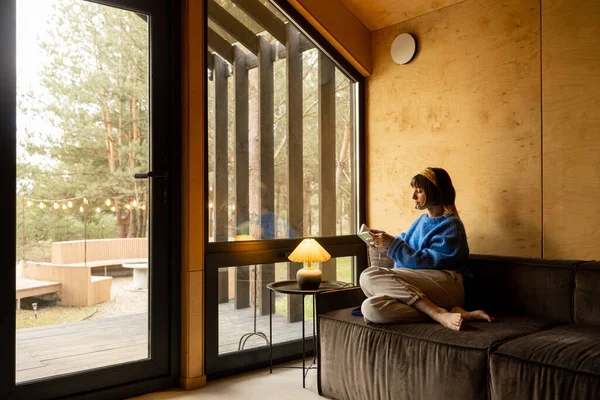 Kobieta Czyta Książkę Siedząc Zrelaksowany Kanapie Przy Oknie Drewnianym Domku — Zdjęcie stockowe