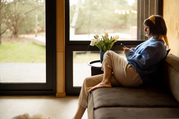 Woman sitting relaxed with a phone on a couch by the window at wooden cottage. Concept of home comfort and leisure time
