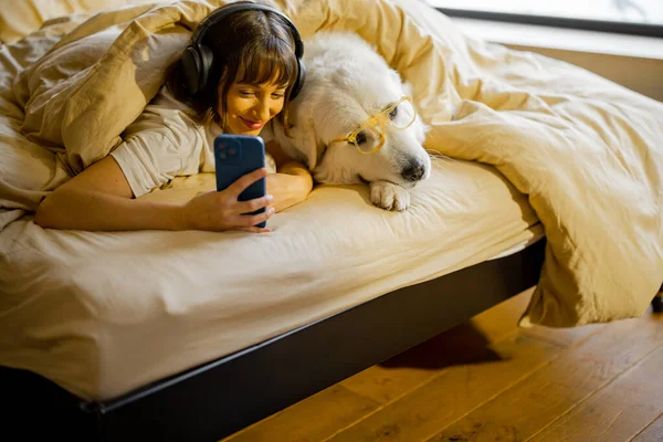 若い女性はベッドの毛布の下で彼女のかわいい犬と横になっている間 自撮り写真を撮るか ビデオ通話を持っています 余暇を過ごし 一緒に楽しみましょう ペットとの友情と居心地の良いコンセプト — ストック写真
