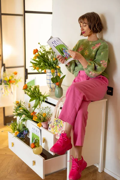 Young woman sits and reads magazine about spring on white commode decorated with bouquets of fresh cutted flowers. Concept of beauty, spring and style