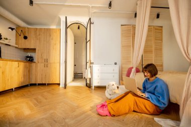 Modern stüdyo dairesindeki bir kadın köpeğiyle yerde otururken dizüstü bilgisayarda çalışıyor. Geniş iç görünüş. Ev konforu ve ev konseptinden çalışma