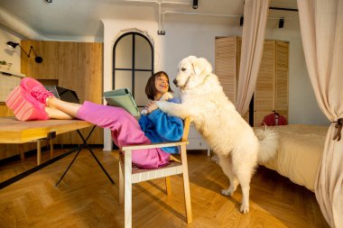 Kadın evde çalışırken masa başında rahatça otururken köpeğiyle oynuyor. Evden çalışmak ve evcil hayvan konseptiyle vakit geçirmek.