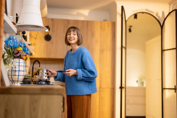 年轻的女人把水壶放在炉子上加热 而站在现代化厨房的内部带着电话 家居休闲和时尚室内装潢的概念 — 图库照片