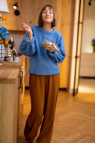 女人在碗里享用着格拉诺拉早餐 站在家里的现代化厨房里 时尚早餐和家庭生活方式的概念 — 图库照片