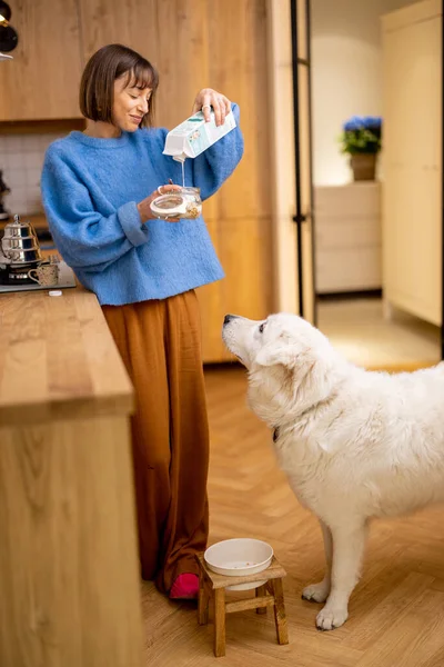 年轻的女人在家里和她可爱的狗一起站在厨房里 用酸奶和麦片做早餐 家庭生活方式和与宠物的友谊概念 — 图库照片