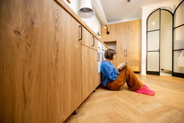 年轻女子坐在新的时尚工作室公寓的厨房地板上 带着一个数码平板电脑 宽的内部视图和复制空间 — 图库照片
