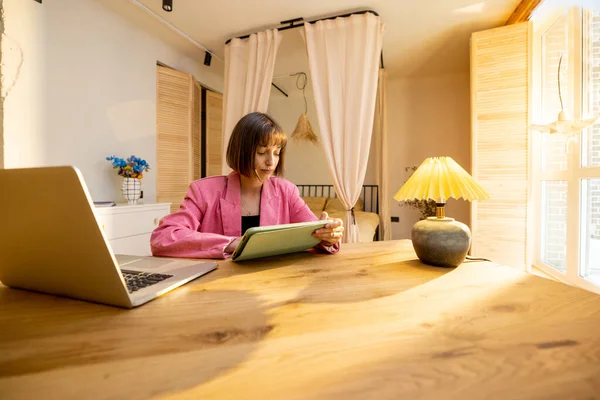 Young business lady in pink suit works on a digital tablet at cozy home office. Concept of freelance and remote work from cozy home atmosphere
