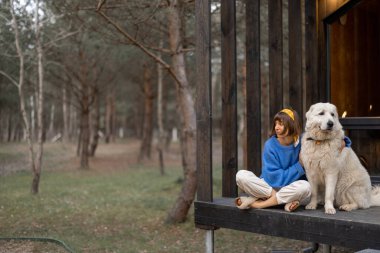 Young woman sits with her dog on porch of a wooden house in pine forest, enjoying nature while resting in cottage at countryside