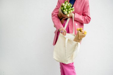 Stylish woman in pink suit takes some magazine from eco handbag and holds flowers on white background. Canvas bag with blank space