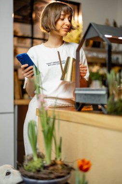 Young woman growing sprouts under artificial light, standing on kitchen with flowers at home. Concept of growing flowers or greenery at home and sustainability