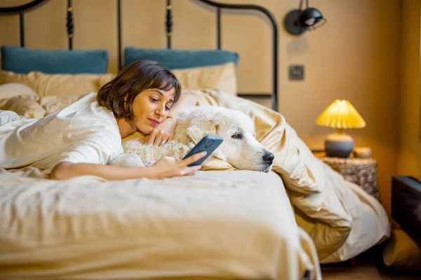 かわいい若い女性は彼女の白い愛らしい犬と抱擁し 自宅でベッドに横たわっている間 電話を使用しています ペットとの友情と家庭の居心地の良さの概念 — ストック写真