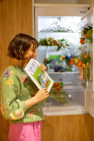 Kadın Içi Taze Çiçeklerle Dolu Buzdolabının Yanında Dururken Dergiyi Isırıyor — Stok fotoğraf