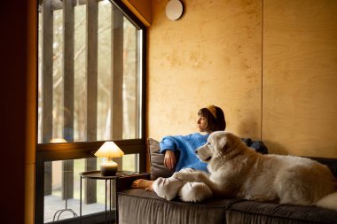 Woman cares her dog while sitting on a couch by the window, spending leisure time together in wooden house on nature