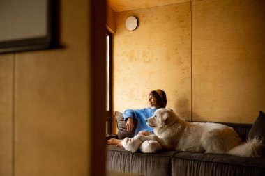 Woman cares her dog while sitting on a couch by the window, spending leisure time together in wooden house on nature