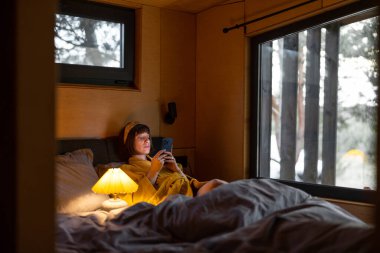 Woman in yellow bathrobe uses a smartphone while lying in bed at tiny bedroom of wooden cabin on nature. Communication online during bedtime and addiction to smartphone