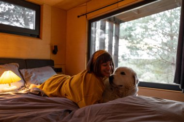 Woman lying with her huge adorable white dog in tiny bedroom while resting in wooden cabin on nature