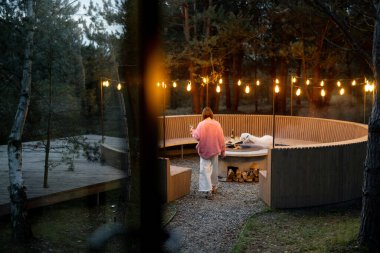 Beautiful lounge area with round bench and bonfire illuminated with garlands near house in pine forest, view from the house