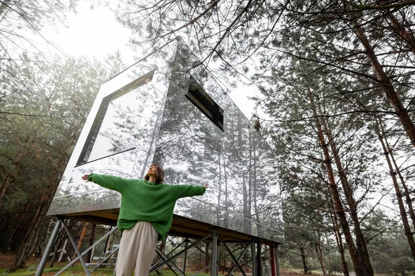 Woman near an invisible house with mirrored walls in pine forest. Connection with nature and sustainability concept. Rest in tiny cabins on nature