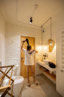 Beyaz ve şık banyoda duş aldıktan sonra saç kurutma makinesiyle saç kurutan genç bir kadın. Sağlık ve ev hayatı kavramı