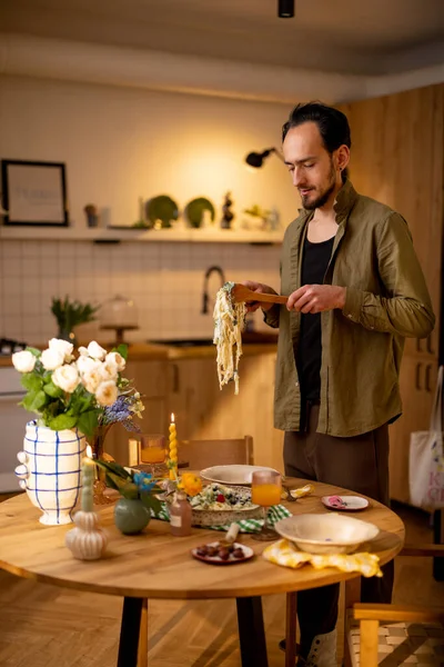 男人在舒适的家里的饭桌上把意大利面放在盘子里 — 图库照片