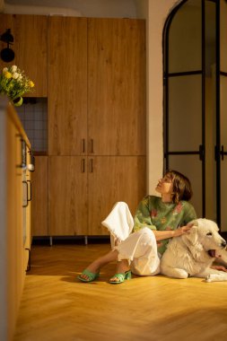 Kadın köpeğiyle birlikte şık bir stüdyo dairede mutfak zemininde oturuyor. Evcil hayvan konsepti ile aile tarzı ve arkadaşlık.